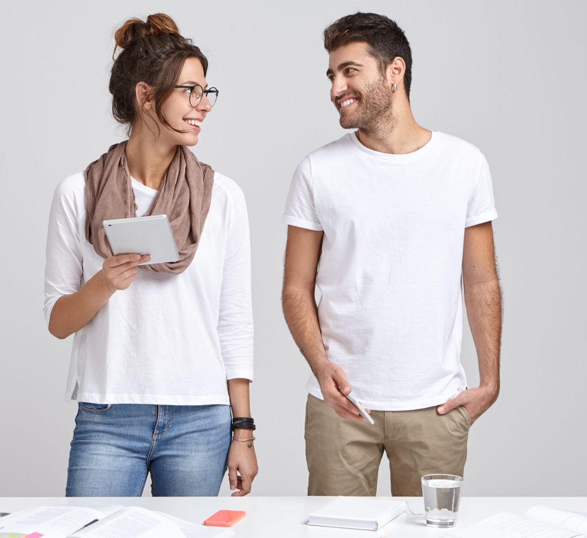 Horizontal shot of two cheerful youngsters groupmates prepare for workshop, hold modern gadgets in hands, look positively at each other, pose near table with magazine, notepad and glass of water